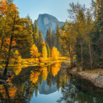 Half Dome reflected in the Merced River in Yosemite National Park, showcasing the clear blue skies and calm waters ideal for fall photography.