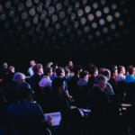 Dozens of people sit in a dark theater taking notes at a corporate conference