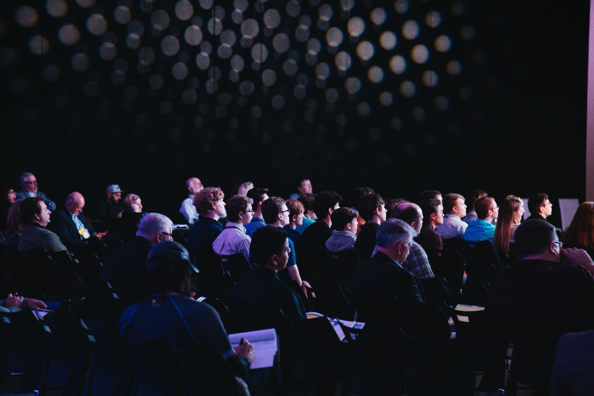 Dozens of people sit in a dark theater taking notes at a corporate conference