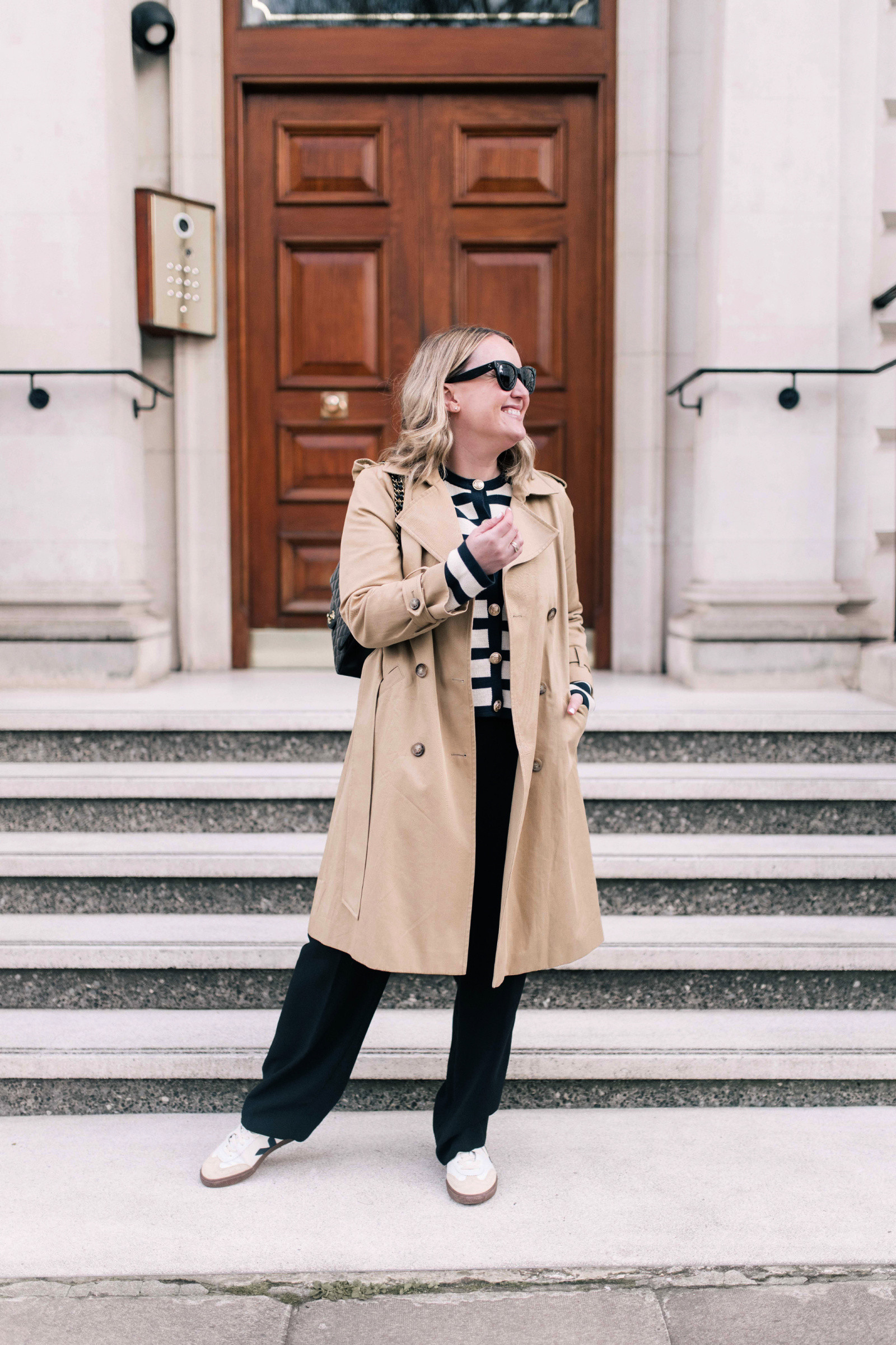Woman wearing a layered travel outfit with a knit sweater jacket, trench coat, and trousers, perfect for navigating different climates during travel.