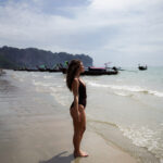 Tourists relaxing on Ao Nang Beach, Krabi, Thailand, with clear blue water and longtail boats in the background.