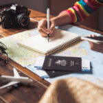 Person making trip notes surrounded by travel items like a world map, passports, and camera