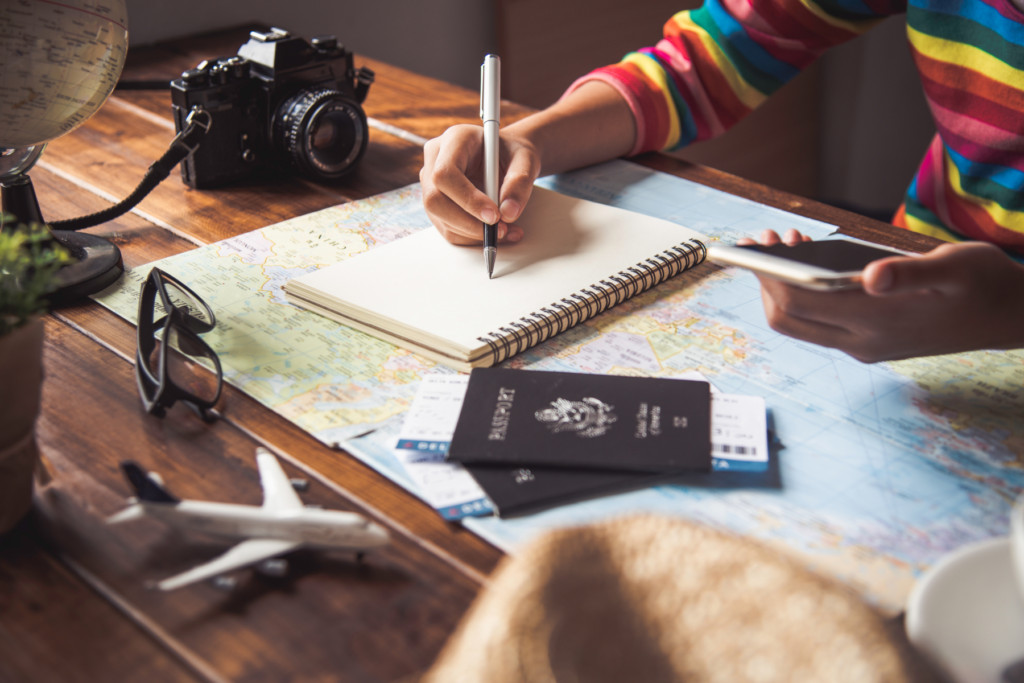 Person making trip notes surrounded by travel items like a world map, passports, and camera