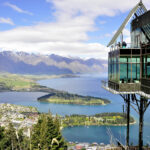 A contemporary glass building perched on a hilltop offers panoramic views of Queenstown's skyline, featuring snow-capped mountains and the expansive Lake Wakatipu with a verdant peninsula extending into its waters.