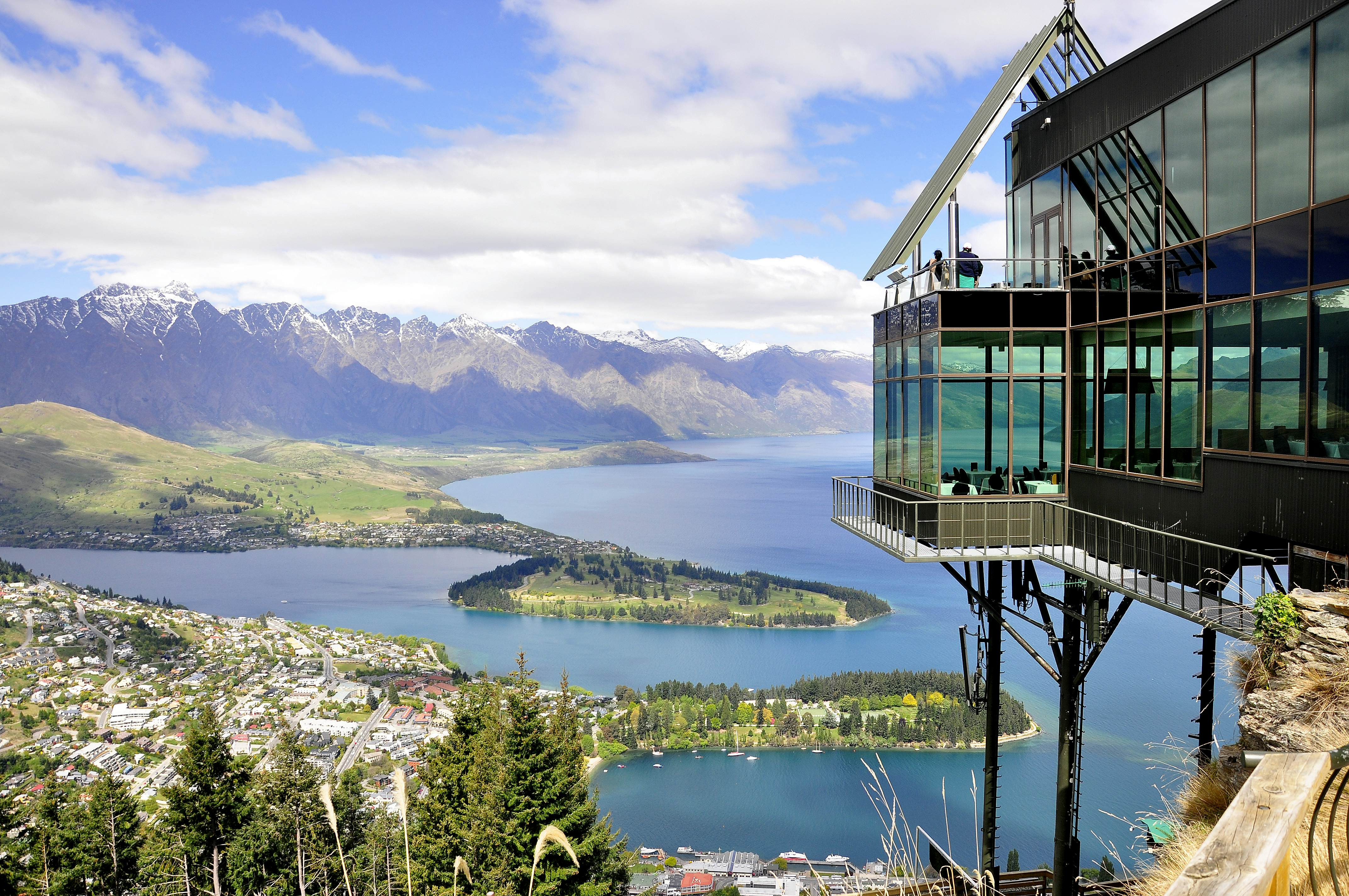 A contemporary glass building perched on a hilltop offers panoramic views of Queenstown's skyline, featuring snow-capped mountains and the expansive Lake Wakatipu with a verdant peninsula extending into its waters.