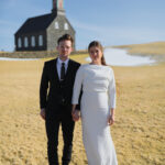 A couple standing in a field in Iceland, surrounded by stunning mountain scenery, showcasing their adventurous spirit and the beauty of their chosen wedding destination.