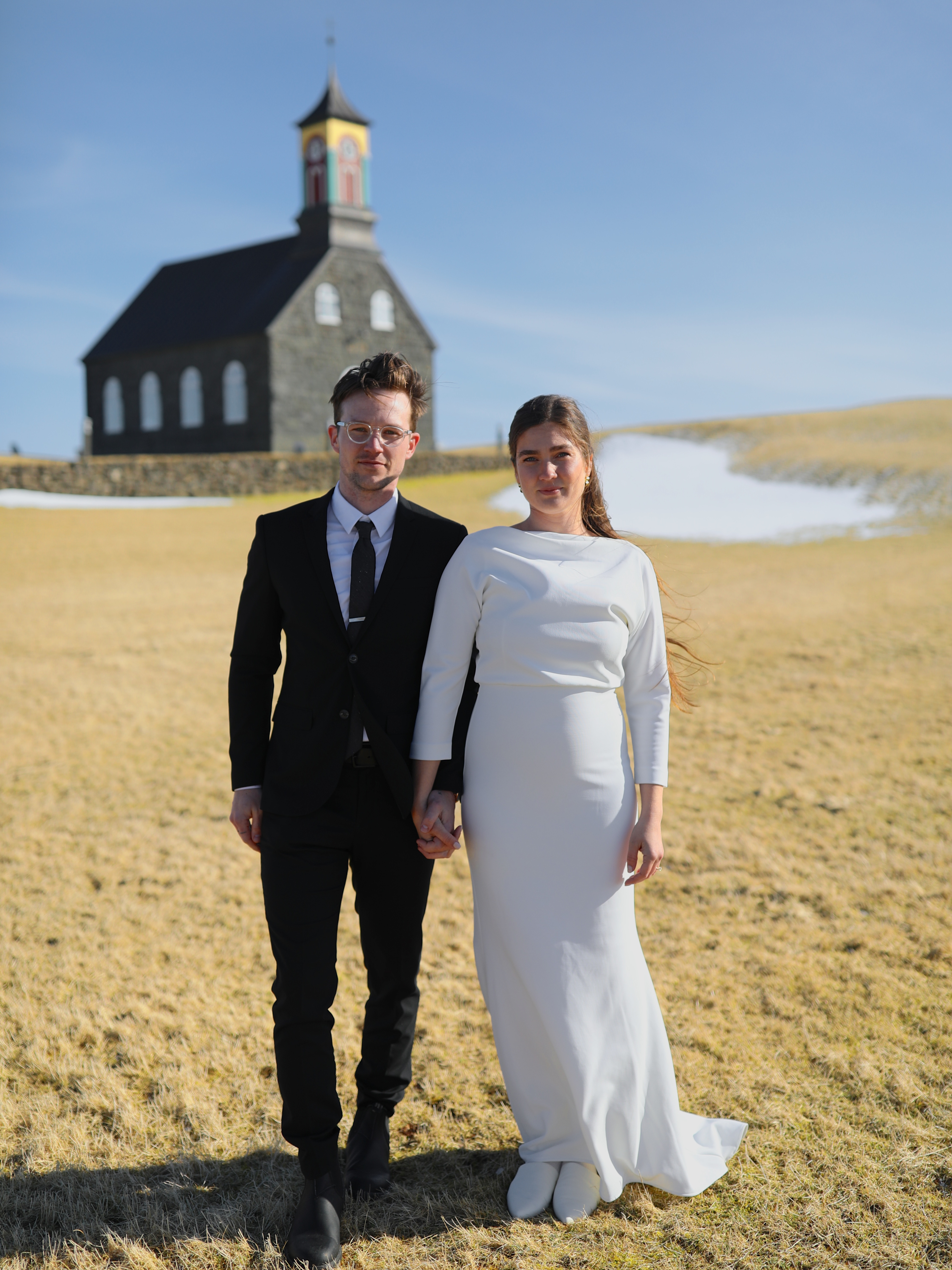 A couple standing in a field in Iceland, surrounded by stunning mountain scenery, showcasing their adventurous spirit and the beauty of their chosen wedding destination.