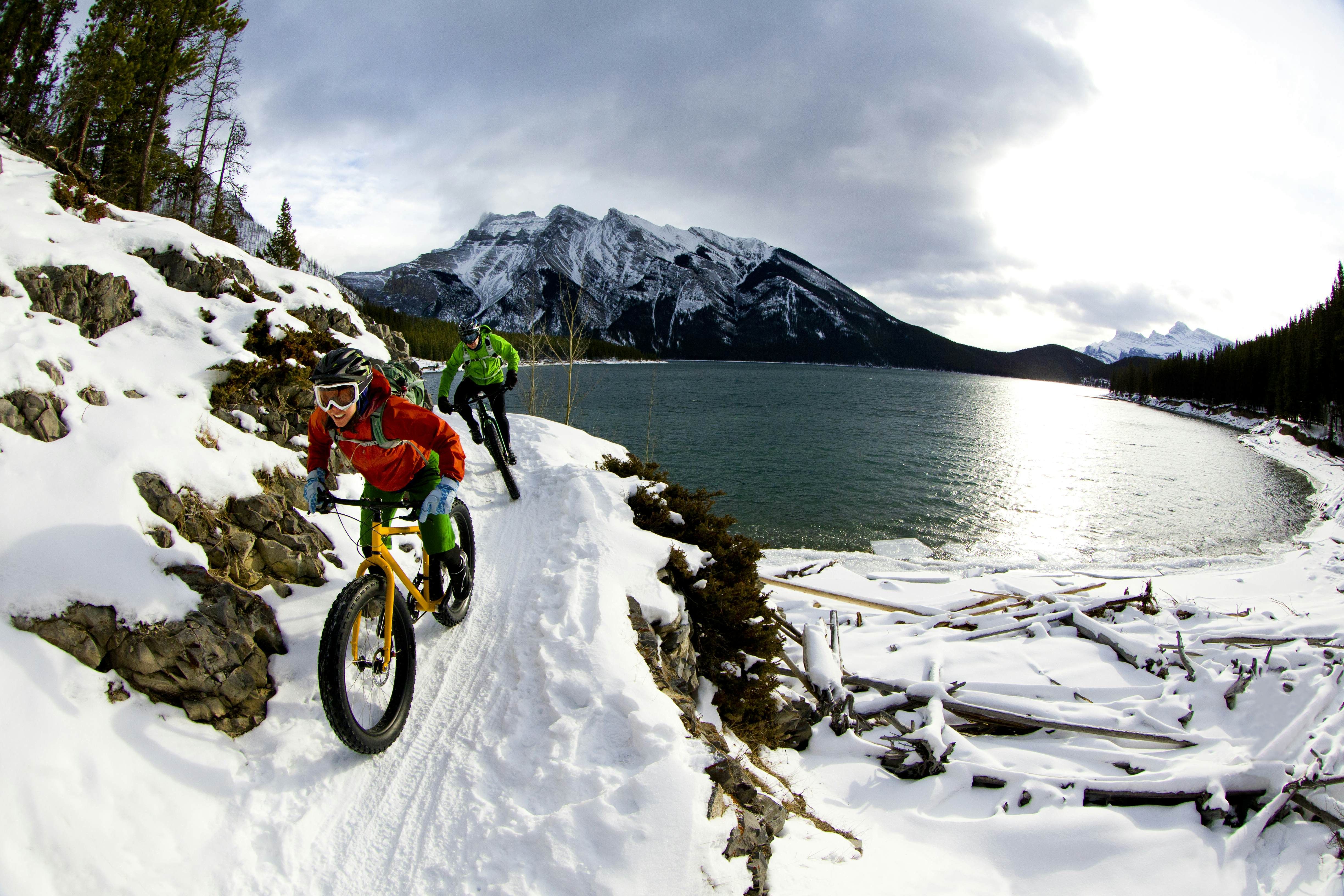 Mountain bikers on a snowy trail by a lake in the Canadian Rockies, highlighting outdoor adventures