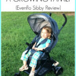 Evenflo Sibby Travel System Review: A parent pushing the stroller with a baby in the car seat and a toddler standing on the ride-along board, highlighting the stroller's functionality for families with multiple young children.