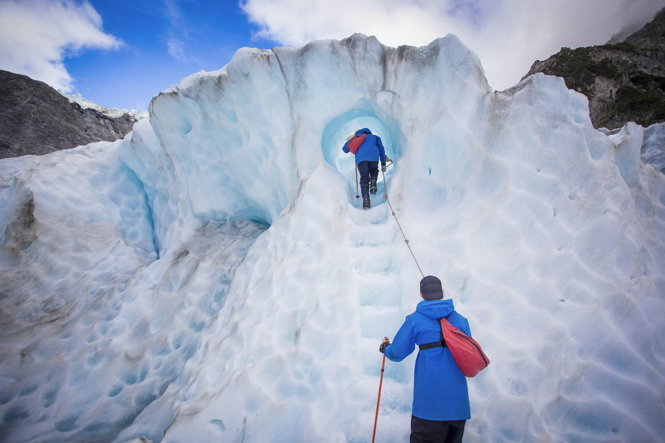 Two hikers, equipped with backpacks and ice axes, ascend a series of ice steps within a glacier tunnel, surrounded by blue glacial ice.