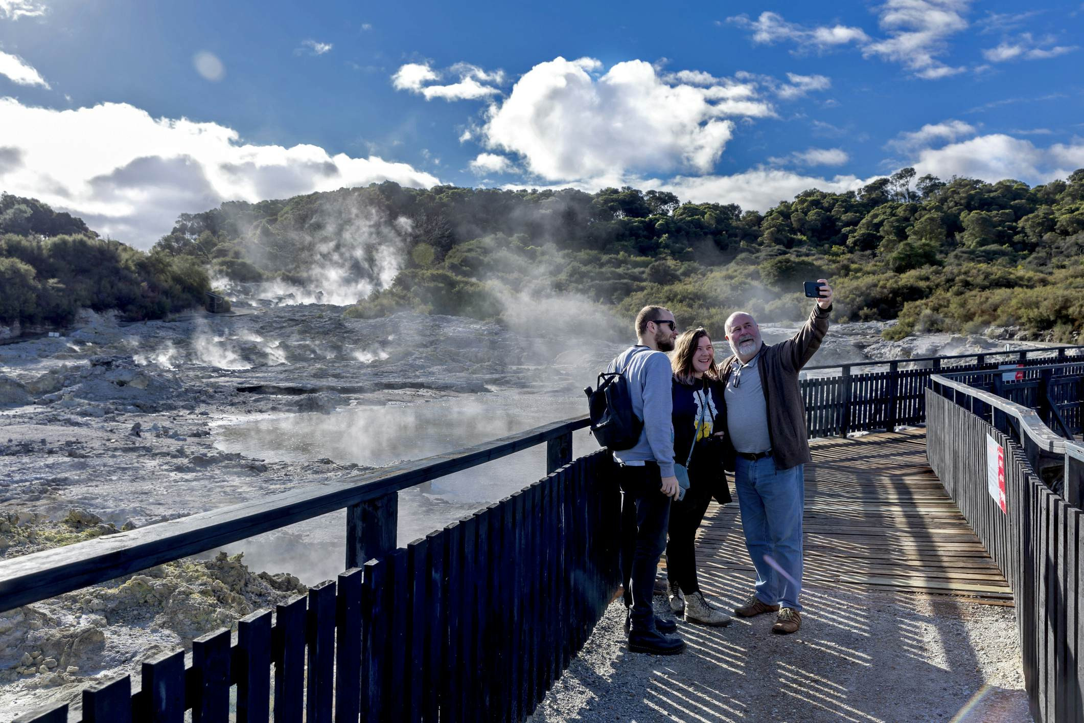 Three individuals of varying ages are positioned on a boardwalk, posing for a selfie with a backdrop of steaming, bubbling geothermal earth.