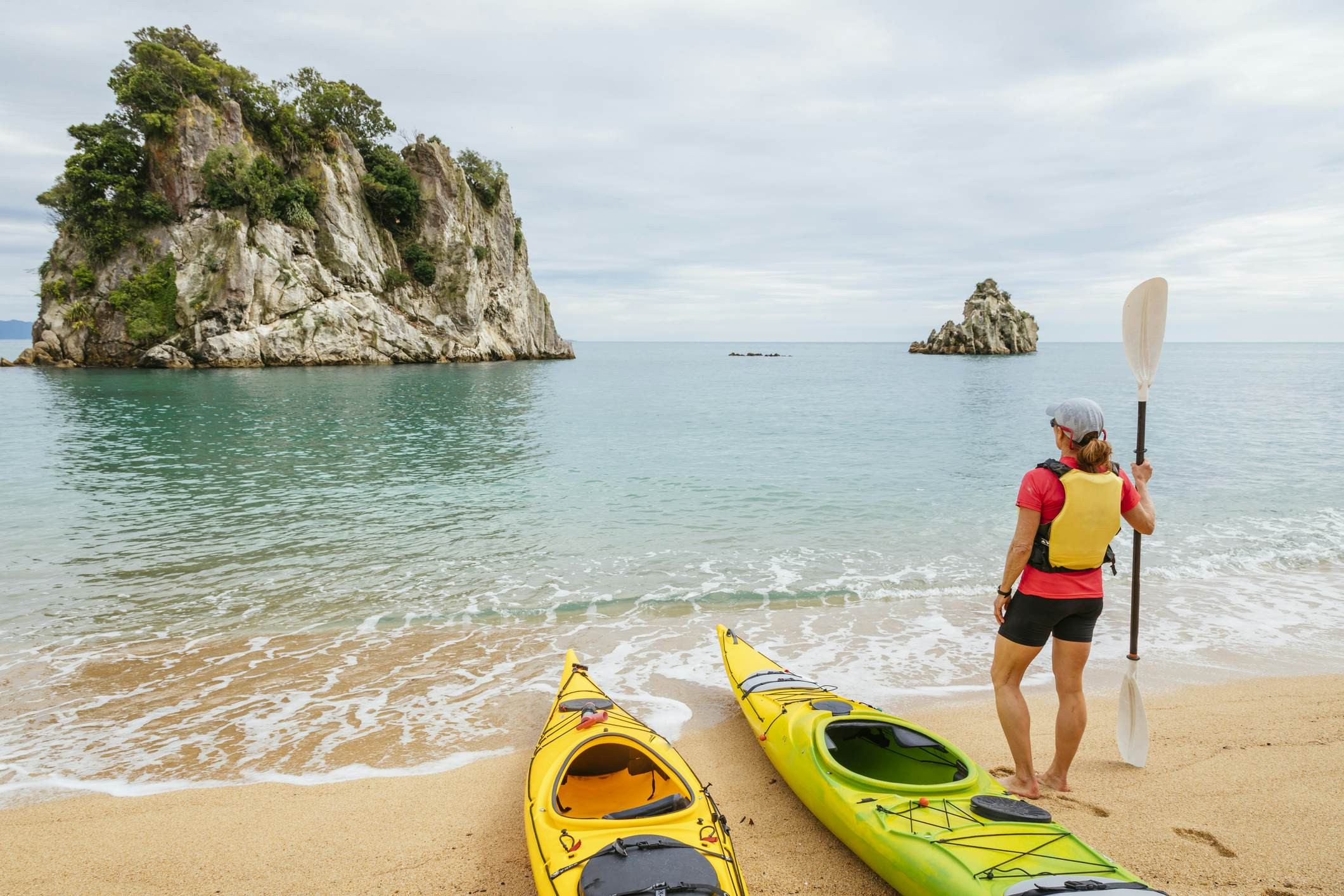 A woman in a blue jacket and black pants stands beside a yellow kayak, gazing out at a prominent rocky outcrop in the ocean, under a slightly overcast sky.