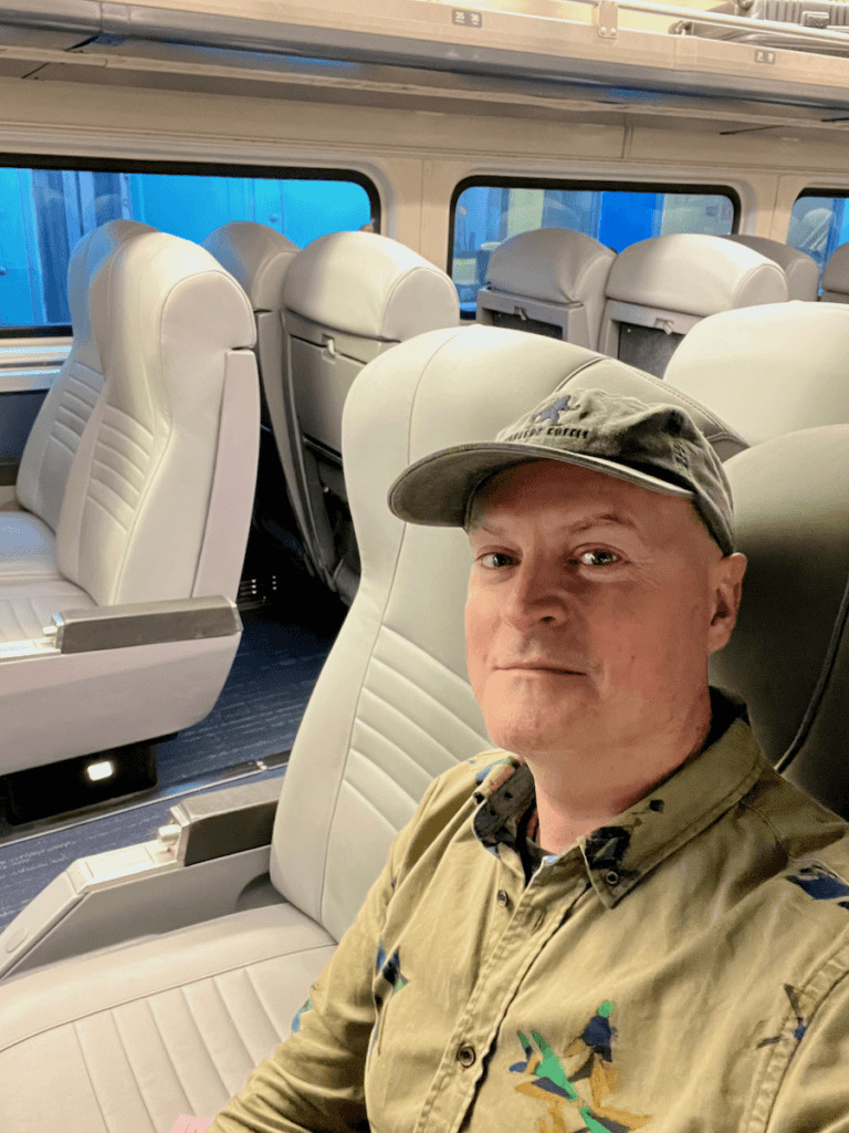Matthew Kessi poses for a selfie in a car on an Amtrak train. He is sitting in plush gray leather seat with others visible behind him. He