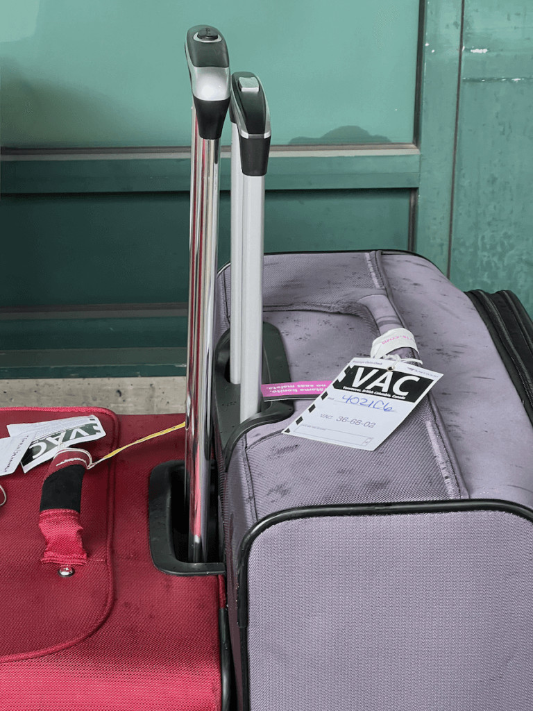 Two pieces of baggage sit back to back, one is red and the other is purple. They have the pull handles up, ready to roll. The luggage tags on both say VAC for Vancouver Canada.