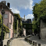 A winding street in Montmartre, Paris, with a stone wall on the right and the famous pink house covered in ivy on the left