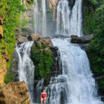 A woman wearing a red two-piece swim suit in posing on a boulder in front of a tall two-tiered waterfall