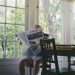 A person sitting at a table with a laptop and notebooks, seemingly working on writing, with a cup of coffee and a magazine visible, representing the work of a travel writer.