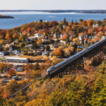Aerial view of a Canadian train gracefully crossing a high trestle bridge amidst a charming waterfront town surrounded by lush green trees