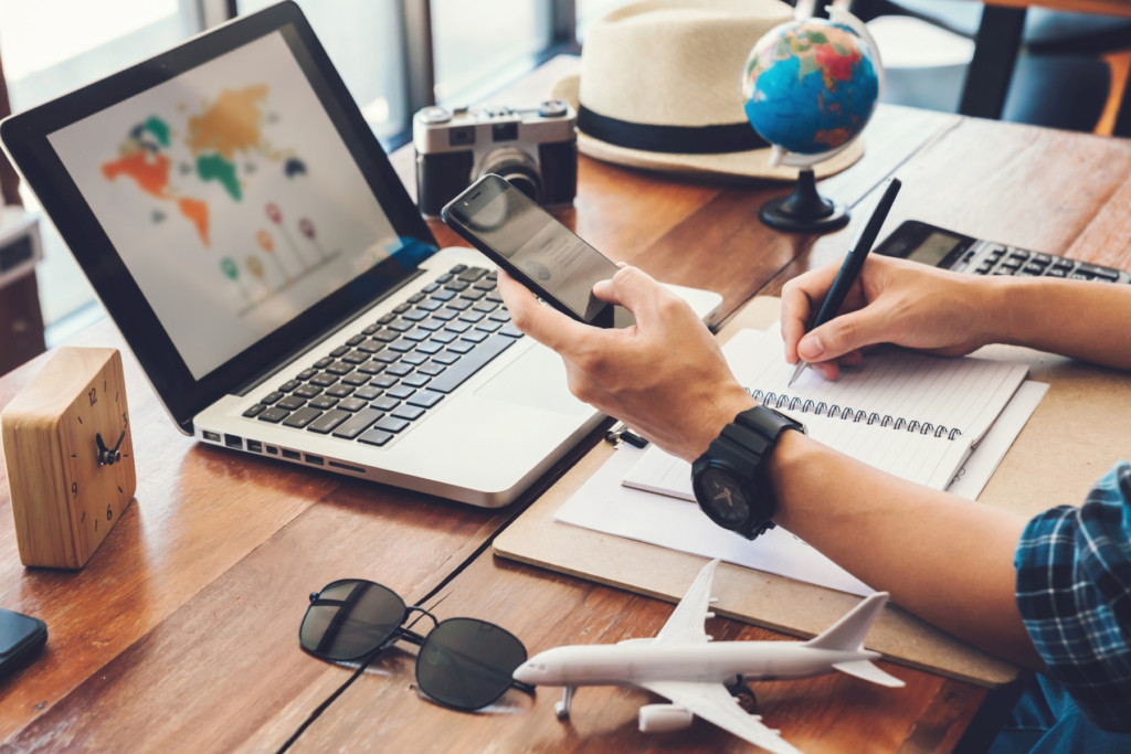 Person holding phone and looking at laptop while making travel notes