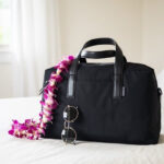 Away The Everywhere Bag on bed with sunglasses resting against the bag, a stylish and practical travel bag for your essentials.